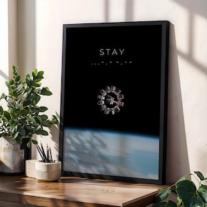 Può includere: Un poster verticale incorniciato con sfondo nero e la parola STAY in lettere maiuscole bianche nella parte superiore. Sotto il testo si trova una piccola illustrazione dettagliata di una stazione spaziale circolare in orbita sopra un pianeta blu.