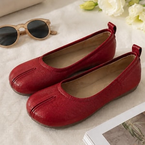 Puede incluir: Un par de bailarinas de cuero rojo oscuro con un diseño distintivo de punta dividida en la parte delantera, una línea superior de corte bajo y una pequeña lengüeta en el talón. Los zapatos tienen un acabado liso y ligeramente texturizado.