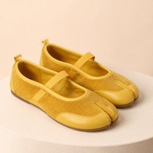 Op de afbeelding: Een paar mosterdgele flats met gespleten neus, bestaande uit een combinatie van glad leer en su&egrave;de met textuur. Deze schoenen zijn voorzien van een bandje over de wreef, een hiellus en een platte zool met een duidelijke scheiding bij de tenen.