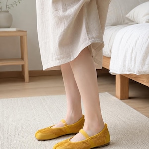 Op de afbeelding: Een paar mosterdgele schoenen met gespleten teen, voorzien van een voorpaneel met su&egrave;de-achtige textuur, een gladde, leerachtige neus en een dun elastisch bandje over de wreef. De schoenen hebben een platte zool en een opvallend splitontwerp.