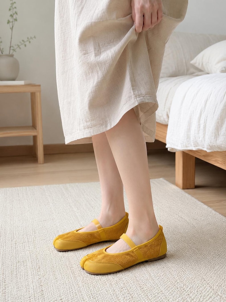 Op de afbeelding: Een paar mosterdgele schoenen met gespleten teen, voorzien van een voorpaneel met su&egrave;de-achtige textuur, een gladde, leerachtige neus en een dun elastisch bandje over de wreef. De schoenen hebben een platte zool en een opvallend splitontwerp.
