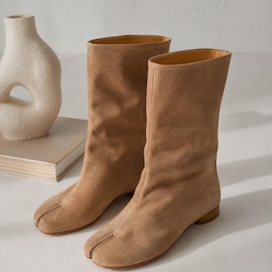 May include: A pair of tan suede mid-calf boots featuring a distinct split-toe design, a flat sole, and a low heel. The boots have a soft, textured exterior with a simple pull-on construction and a smooth interior lining visible at the top opening.