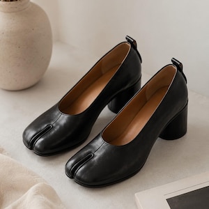 May include: A pair of black leather shoes with a split-toe design, featuring a low block heel and a small pull tab at the back of the heel collar. The shoes have a smooth finish, a rounded topline, and a tan interior lining visible at the opening.