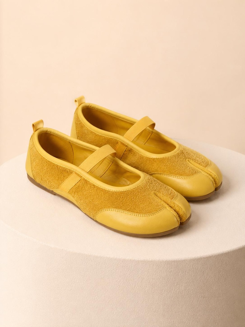 Op de afbeelding: Een paar mosterdgele flats met gespleten neus, bestaande uit een combinatie van glad leer en su&egrave;de met textuur. Deze schoenen zijn voorzien van een bandje over de wreef, een hiellus en een platte zool met een duidelijke scheiding bij de tenen.