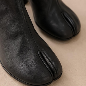 K&ouml;nnte beinhalten: Ein Paar schwarze Lederstiefeletten mit einem markanten Design mit geteilter Zehenpartie. Die Stiefel haben eine glatte, matte Oberfl&auml;che mit einer vertikalen Naht, die &uuml;ber die Mitte des Vorderblatts bis zur charakteristischen Zehenspalte verl&auml;uft.