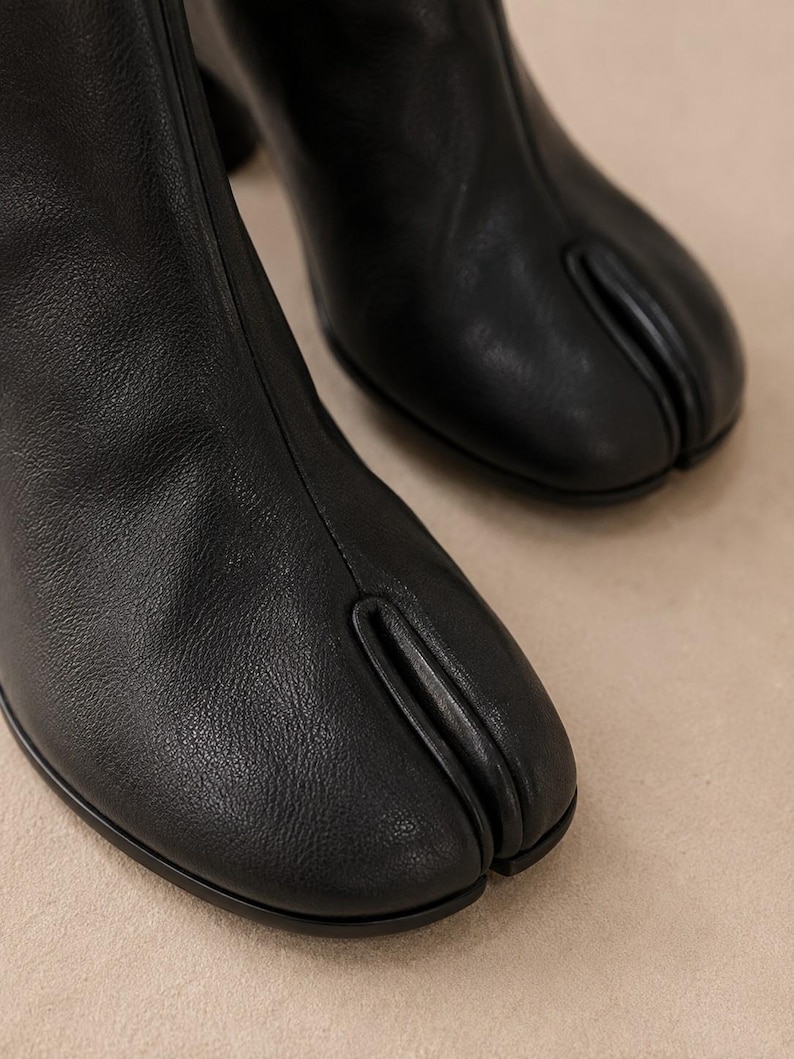 K&ouml;nnte beinhalten: Ein Paar schwarze Lederstiefeletten mit einem markanten Design mit geteilter Zehenpartie. Die Stiefel haben eine glatte, matte Oberfl&auml;che mit einer vertikalen Naht, die &uuml;ber die Mitte des Vorderblatts bis zur charakteristischen Zehenspalte verl&auml;uft.