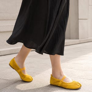 Op de afbeelding: Een paar felgele ballerina's met gespleten teen, een afgeronde neus, een dun elastisch bandje over de wreef en een platte zool. De schoenen worden gedragen met een zwarte rok die tot halverwege de kuit valt en het schoeisel accentueert.