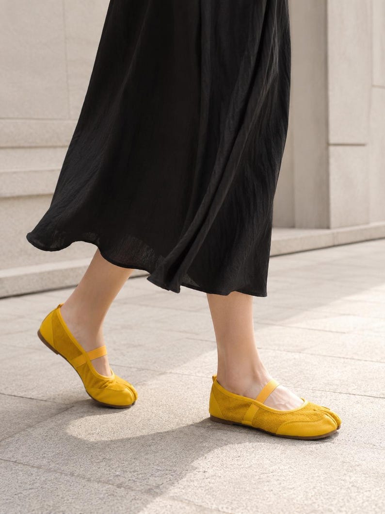 Op de afbeelding: Een paar felgele ballerina's met gespleten teen, een afgeronde neus, een dun elastisch bandje over de wreef en een platte zool. De schoenen worden gedragen met een zwarte rok die tot halverwege de kuit valt en het schoeisel accentueert.
