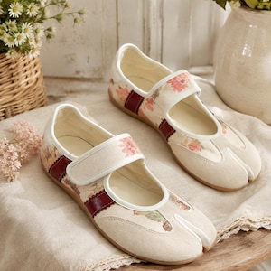 May include: A pair of split-toe shoes featuring a cream-colored fabric upper with a floral pattern, accented by a dark red strap across the midfoot. The shoes have a hook-and-loop closure, a white trim, and a flat tan sole, showcasing a distinct toe separation.