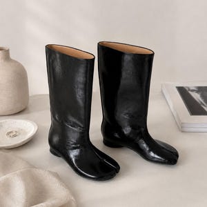 May include: A pair of black mid-calf boots with a glossy finish and a distinct split-toe design. The boots feature a smooth, structured shaft, a low heel, and a rounded silhouette, showcasing a polished appearance with visible natural leather creasing.