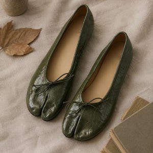 May include: A pair of olive green split-toe ballet flats with a glossy, crinkled texture. Each shoe features a thin, matching green bow detail at the center of the vamp. The design includes a distinct separation between the big toe and other toes.