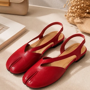 May include: A pair of bright red flat shoes featuring a split-toe design, a slingback strap, and a V-shaped cutout on the upper. The shoes have a smooth finish, a tan interior lining, and a low heel, presenting a distinct silhouette.