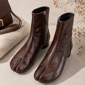 May include: A pair of dark brown ankle boots with a glossy, crinkled patent leather finish. These boots feature a distinctive split-toe design, a vertical seam running up the front, and a side zipper closure with a low, flat heel for stability.