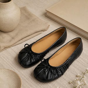 May include: A pair of black ballet flats featuring a soft, gathered leather upper with a small decorative bow at the rounded toe. The interior of the shoes is lined in a tan color, and the design includes a low-profile sole for a flat silhouette.