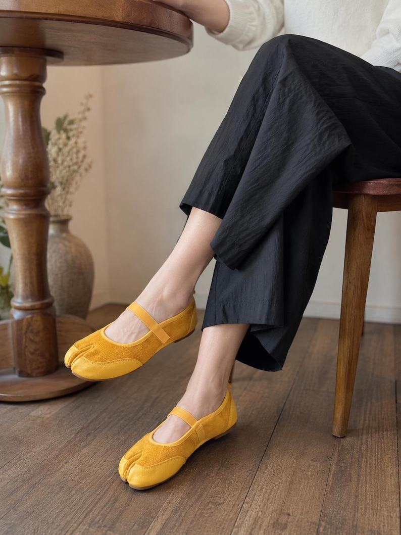 Op de afbeelding: Een paar felgele schoenen met een gespleten teen, voorzien van een textuur die lijkt op su&egrave;de aan de voorzijde en een glad materiaal aan de achterzijde. Deze schoenen hebben een dun bandje over de wreef en een platte zool.