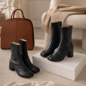 May include: A pair of black leather ankle boots featuring a distinct split-toe design and a block heel. The boots have a smooth finish, a mid-calf height, and a structured silhouette. The design includes a vertical seam running up the front center.