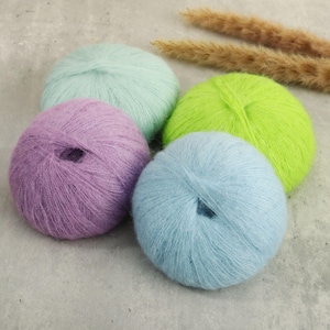 Peut inclure: Quatre pelotes de laine duveteuse de forme ronde dans des tons pastel bleu clair, lavande, vert menthe et vert citron. Le fil présente une texture douce et brossée, disposé en groupe. Chaque pelote est sphérique et faite de fibres fines.