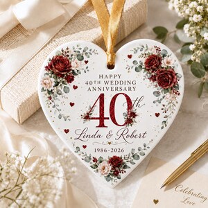 May include: A heart-shaped white ceramic ornament featuring a gold ribbon hanger. The design includes dark red roses, eucalyptus leaves, and small red hearts. Text reads: Happy 40th Wedding Anniversary, 40th, Linda & Robert, and 1986-2026.