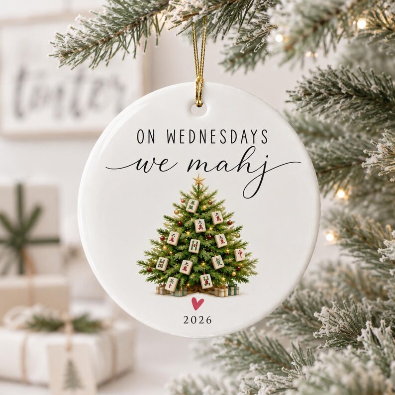 May include: A round white ceramic Christmas ornament featuring the text ON WEDNESDAYS we mahj, a green Christmas tree decorated with Mahjong tiles, a small red heart, and the year 2026 printed in black, suspended by a gold string.