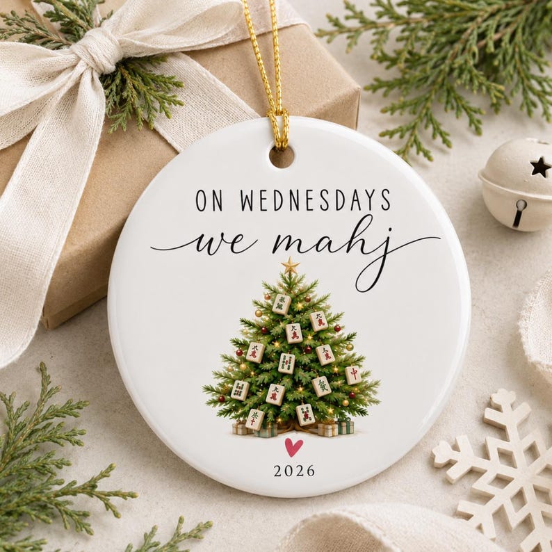 May include: A round white ceramic Christmas ornament featuring a green pine tree decorated with mahjong tiles. The text reads ON WEDNESDAYS we mahj above the tree and 2026 with a small red heart below. A gold string is attached for hanging.