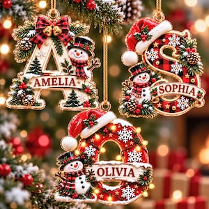 May include: Three hanging Christmas ornaments shaped as letters A, S, and O. Each letter features a snowman, pine branches, pinecones, and a banner with the names AMELIA, SOPHIA, and OLIVIA. The ornaments are decorated with red and black plaid patterns, white snowflakes, and small gold bells.