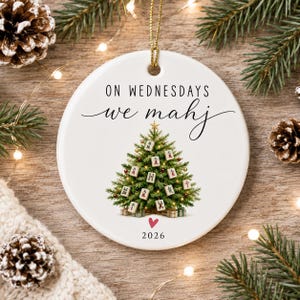 May include: A round white ceramic ornament featuring the black text ON WEDNESDAYS we mahj, a green Christmas tree decorated with Mahjong tiles, a small pink heart, and the year 2026 printed at the bottom, suspended by a gold string.