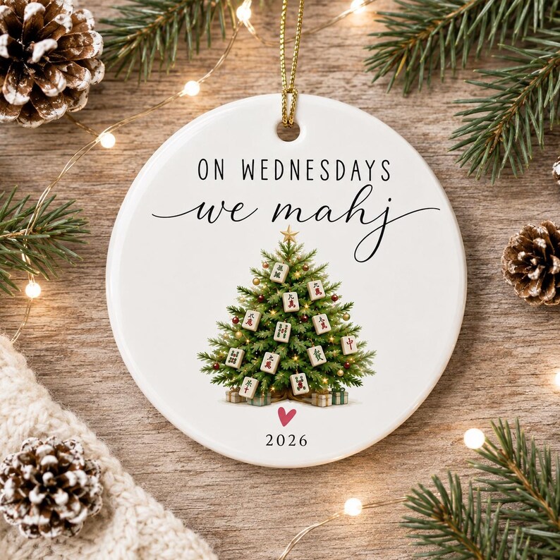 May include: A round white ceramic ornament featuring the black text ON WEDNESDAYS we mahj, a green Christmas tree decorated with Mahjong tiles, a small pink heart, and the year 2026 printed at the bottom, suspended by a gold string.