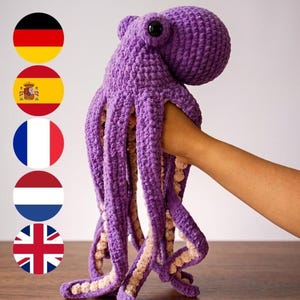 Könnte beinhalten: Ein gehäkeltes Oktopus-Stofftier in Violett mit einem abgerundeten Kopf, einem glatten schwarzen Kunststoffauge und acht langen, herabhängenden Tentakeln mit einer hellcremefarbenen, strukturierten Unterseite. Das Garn wirkt weich und flauschig.