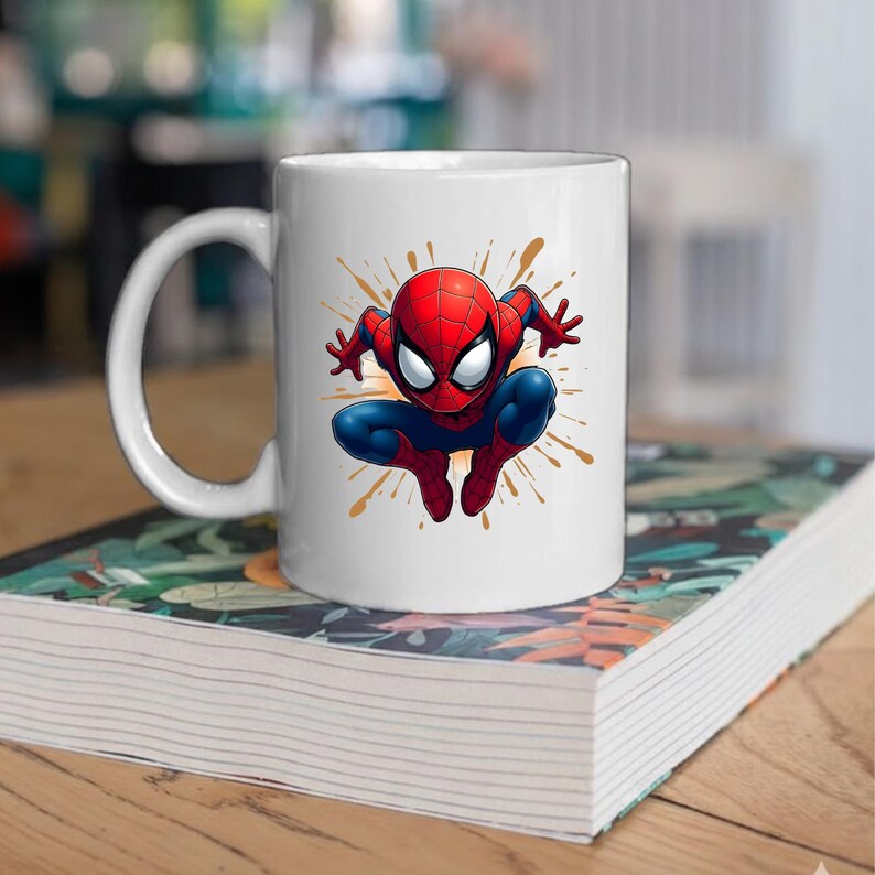 May include: A white ceramic mug featuring a colorful graphic of a red and blue superhero character in a crouching pose, surrounded by orange and brown paint splatter effects. The mug has a smooth finish and a curved handle on the left side.