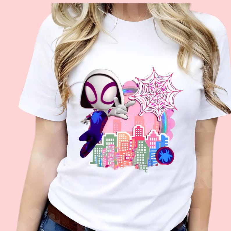 May include: A white short-sleeved t-shirt featuring a graphic print of a stylized character with a white hood and purple mask, a pink spider web, a colorful city skyline, and a small circular emblem with a blue spider icon on a dark background.