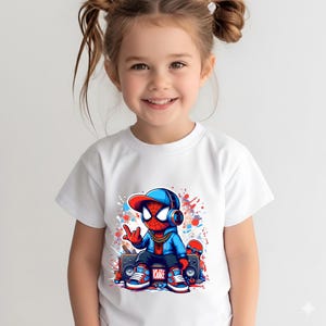 May include: A white t-shirt featuring a graphic print of a stylized character wearing a blue hoodie, a red and blue baseball cap, headphones, and a gold chain. The character sits on a boombox with a skateboard nearby, surrounded by paint splatters.
