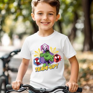 May include: A white short-sleeved t-shirt featuring a graphic print of the Hulk, Spider-Man, and Ghost-Spider inside hexagonal frames. The design includes yellow spiderwebs and the text "TEAM-UP!" in bold yellow letters with a blue outline.