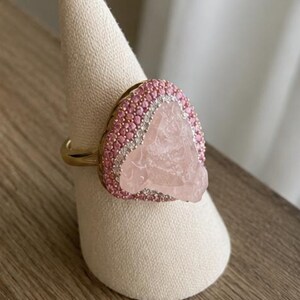 May include: A gold ring featuring a large, irregular, pale pink translucent stone at the center. The stone is surrounded by a border of small, round, bright pink stones, followed by an outer halo of tiny, clear, sparkling white stones.