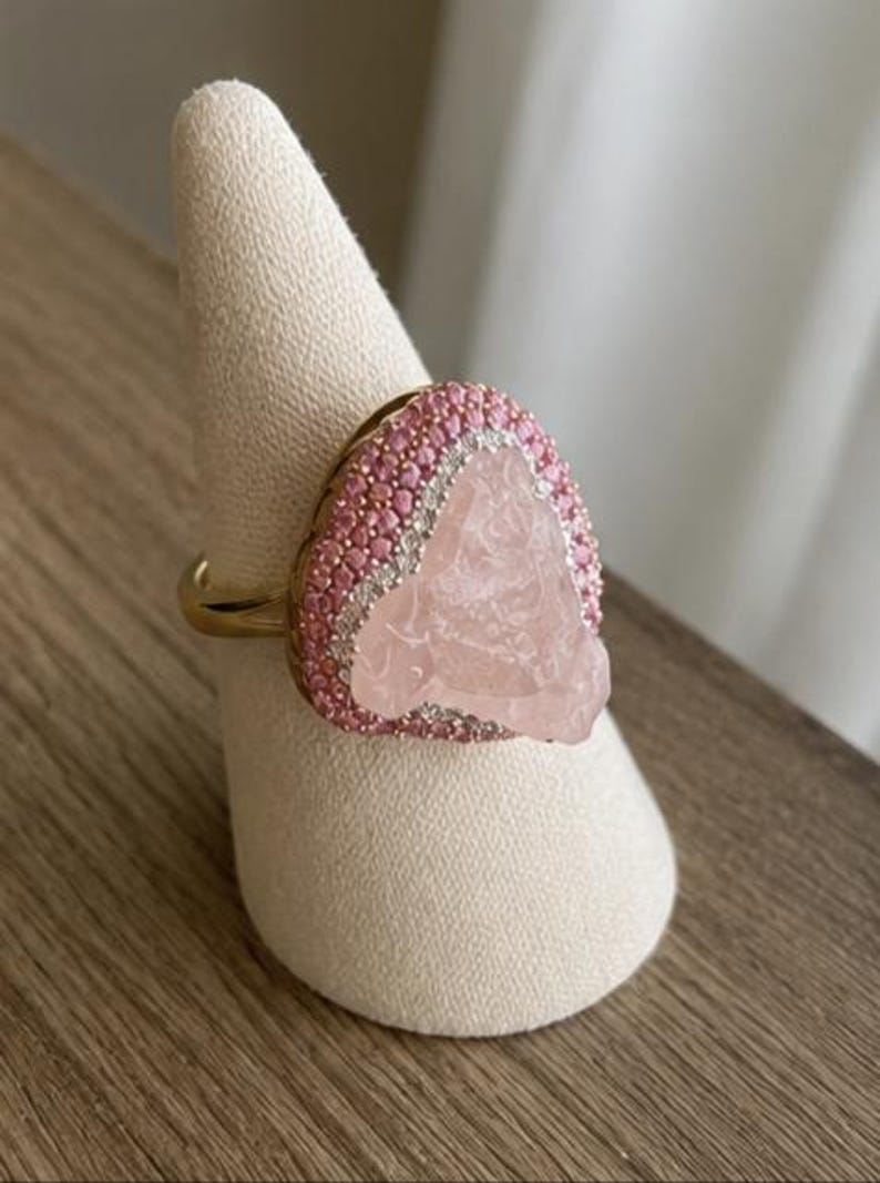 May include: A gold ring featuring a large, irregular, pale pink translucent stone at the center. The stone is surrounded by a border of small, round, bright pink stones, followed by an outer halo of tiny, clear, sparkling white stones.
