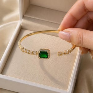 May include: A gold-toned open cuff bracelet featuring a central square-cut emerald green gemstone surrounded by a halo of small clear stones, with additional clear baguette-cut stones set along the band on both sides of the central setting.