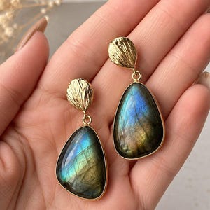 May include: A pair of drop earrings featuring teardrop-shaped labradorite gemstones with iridescent blue and green flashes, set in gold-toned metal frames with textured, shell-like gold-toned studs at the top of each earring.