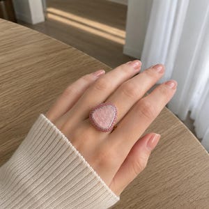 May include: A ring featuring a large, irregular, light pink stone centerpiece. The stone is surrounded by a double border consisting of an inner row of small white stones and an outer row of small pink stones, set on a gold-toned metal band.