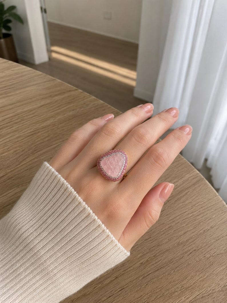 May include: A ring featuring a large, irregular, light pink stone centerpiece. The stone is surrounded by a double border consisting of an inner row of small white stones and an outer row of small pink stones, set on a gold-toned metal band.