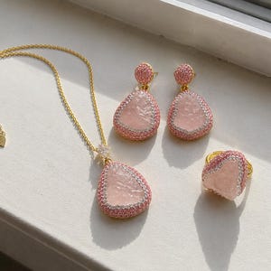 May include: A jewelry set consisting of a gold chain necklace with a teardrop-shaped pink stone pendant, matching teardrop-shaped drop earrings, and a ring. Each piece features a textured pink center stone bordered by small pink crystals.