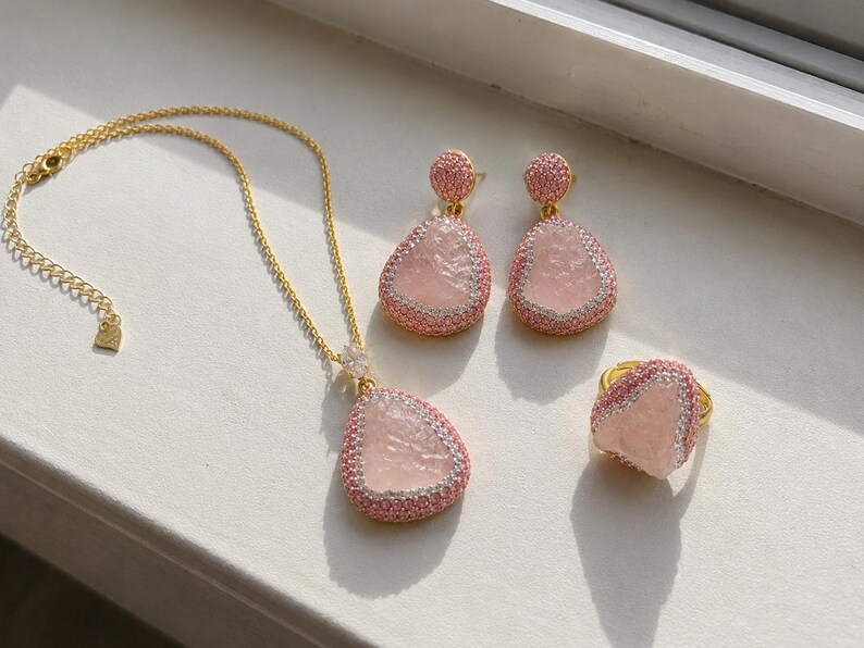 May include: A jewelry set consisting of a gold chain necklace with a teardrop-shaped pink stone pendant, matching teardrop-shaped drop earrings, and a ring. Each piece features a textured pink center stone bordered by small pink crystals.