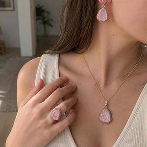 May include: A jewelry set consisting of a gold-tone necklace with a teardrop-shaped pink stone pendant, matching teardrop-shaped drop earrings, and a matching ring. Each piece features a central pink stone surrounded by a border of small pink crystals.