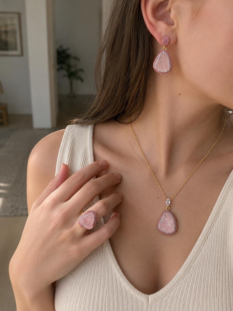 May include: A jewelry set consisting of a gold-tone necklace with a teardrop-shaped pink stone pendant, matching teardrop-shaped drop earrings, and a matching ring. Each piece features a central pink stone surrounded by a border of small pink crystals.