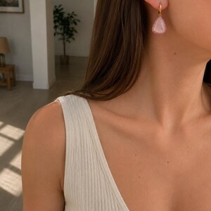 May include: A gold hoop earring featuring a teardrop-shaped pendant with a textured, light pink center and a border of small, sparkling pink stones. The earring hangs from a simple gold hoop, creating a delicate and refined piece of jewelry.