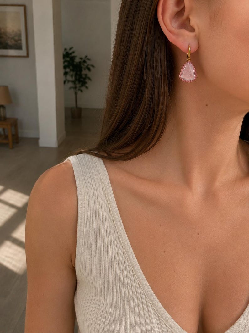 May include: A gold hoop earring featuring a teardrop-shaped pendant with a textured, light pink center and a border of small, sparkling pink stones. The earring hangs from a simple gold hoop, creating a delicate and refined piece of jewelry.