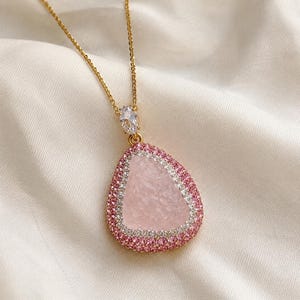 May include: A gold-tone necklace featuring a teardrop-shaped pendant with a textured light pink center stone. The pendant is bordered by a row of clear white stones and an outer row of small pink stones, with a single clear oval stone at the top.