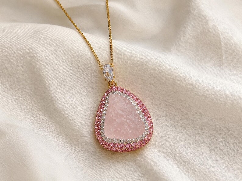 May include: A gold-tone necklace featuring a teardrop-shaped pendant with a textured light pink center stone. The pendant is bordered by a row of clear white stones and an outer row of small pink stones, with a single clear oval stone at the top.