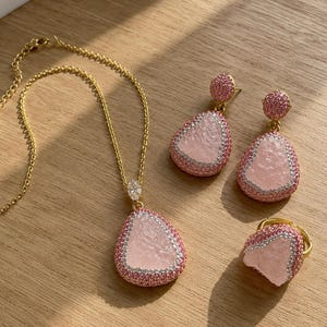 May include: A gold-tone jewelry set consisting of a necklace, a pair of drop earrings, and a ring. Each piece features a central pale pink textured stone framed by small pink and clear crystals, with the necklace chain including a lobster clasp.