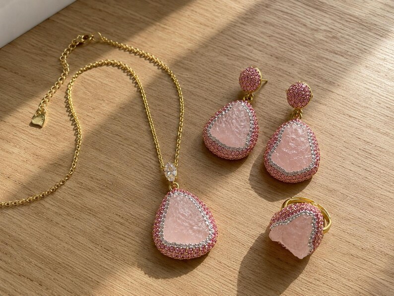 May include: A gold-tone jewelry set consisting of a necklace, a pair of drop earrings, and a ring. Each piece features a central pale pink textured stone framed by small pink and clear crystals, with the necklace chain including a lobster clasp.
