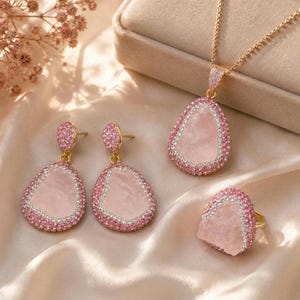 May include: A jewelry set consisting of a necklace, a pair of drop earrings, and a ring. Each piece features a central pale pink stone with an irregular shape, surrounded by a border of small pink and clear crystals set in gold-toned metal.
