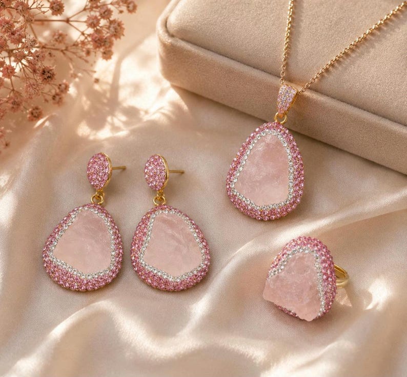 May include: A jewelry set consisting of a necklace, a pair of drop earrings, and a ring. Each piece features a central pale pink stone with an irregular shape, surrounded by a border of small pink and clear crystals set in gold-toned metal.